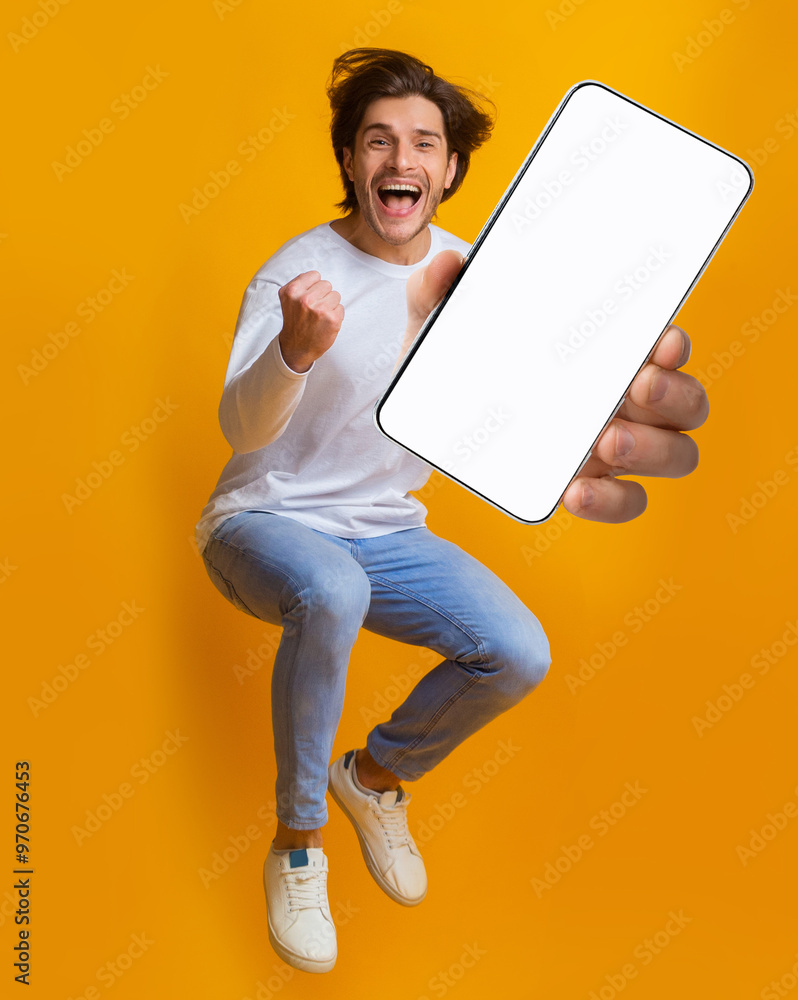 Poster new awesome mobile app. jumping emotional young guy showing smartphone with blank screen on orange s