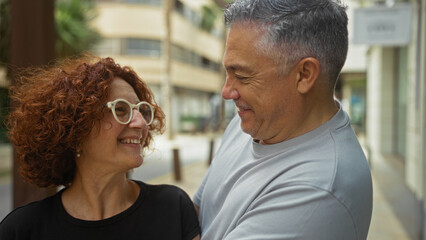 Middle-aged couple smiling warmly at each other while standing on a city street outdoors, conveying love and companionship in an urban setting.