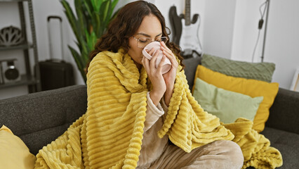 A woman wrapped in a yellow blanket sips tea on a couch indoors, evoking comfort and relaxation at home.