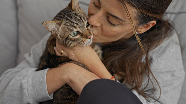 A young hispanic woman cuddles and kisses her pet cat in the living room, displaying affection and warmth while indoors with her feline companion.