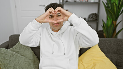 A cheerful man gestures a heart with his hands, smiling in a cozy living room setting, showing positivity and comfort.