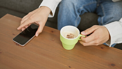 A man in casual attire holding a coffee mug next to a smartphone on a wooden table indoors.