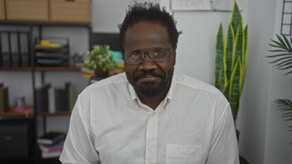 Naklejka premium Handsome african american man with a beard sitting in an office room wearing glasses and a white shirt.