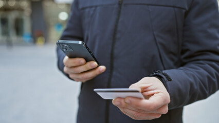 Young man using smartphone and holding credit card on city street, suggesting urban payment or shopping scenario.