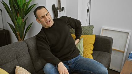 Hispanic man experiencing back pain while sitting on a gray couch in a modern living room.