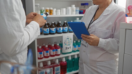Pharmacists working in a pharmacy, with a man reading a medication label and a woman using a digital tablet, standing in front of shelves full of medicines and bottles