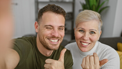 Selfie of a smiling man and woman giving thumbs up in a cozy modern living room.