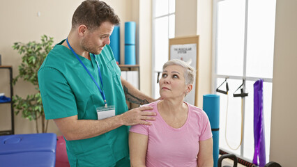 Obraz premium Man nurse in scrubs consults attentive elderly woman patient in bright physiotherapy clinic room.