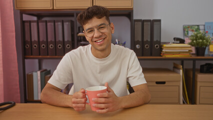 Young hispanic man in an office holding a coffee mug, smiling, and sitting at a desk with organized folders and shelves in the background.