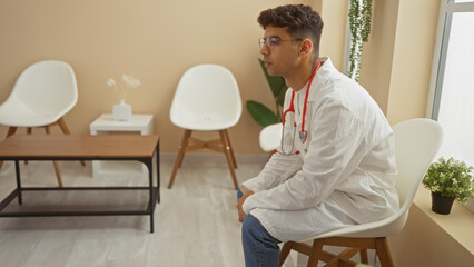 Young, attractive hispanic man in a clinic waiting room wearing a stethoscope and white coat, seated indoors in a modern hospital workplace setting.