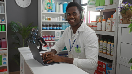 Young man working in a pharmacy store with a laptop, wearing headphones, and smiling, surrounded by shelves of products in an indoor setting