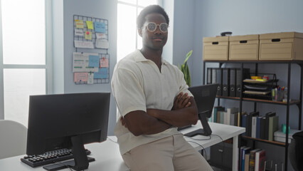 Young, african, american, man, in, office, sits, on, desk, with, arms, crossed, looking, confident, in, workplace, setting