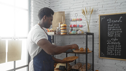 Young african american man arranging pastries in a bakery shop surrounded by a variety of bread and...