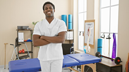 Obraz premium Smiling african american male healthcare professional with arms crossed standing in a modern rehab clinic interior.