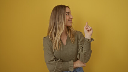 Attractive young blonde caucasian woman posing against an isolated yellow background, pointing upwards with a confident expression, wearing a stylish green blouse and jeans.