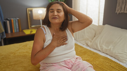 A mature, hispanic woman with short hair wearing pajamas sits on a bed in a cozy bedroom, enjoying...