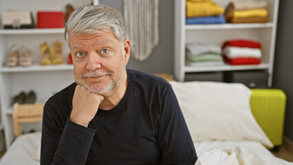 A contemplative mature man with gray hair sitting on a bed in a cozy bedroom, exuding casual...