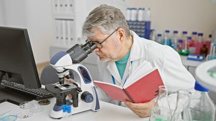 Grey-haired man in lab coat using microscope and holding a notebook in a laboratory setting.