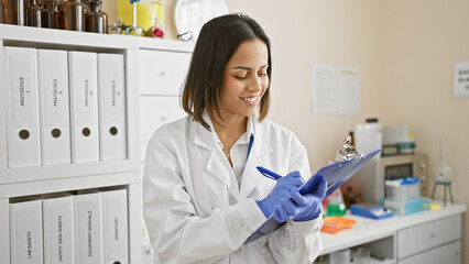 A smiling confident woman scientist in a white lab coat holding a clipboard in a well-organized...