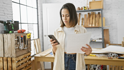 Hispanic woman reads a document while holding smartphone in sunny carpentry workshop