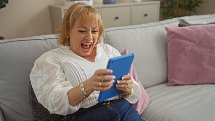 An elderly caucasian woman laughing joyfully while using a blue tablet, sitting comfortably in her living room at home.