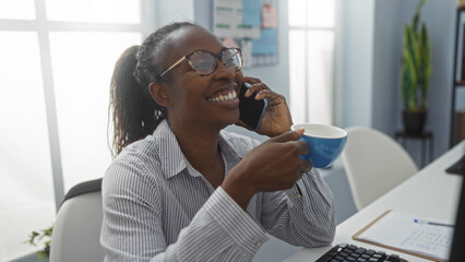 Woman smiling while talking on phone and holding coffee in an office with natural light from large windows