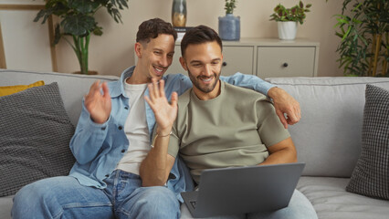 Two men sit on a living room couch, smiling and waving while engaged in a video call at home,...