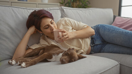 Young caucasian woman relaxing with her pet dog on a couch in the living room, sipping from a cup in a cozy home interior setting.