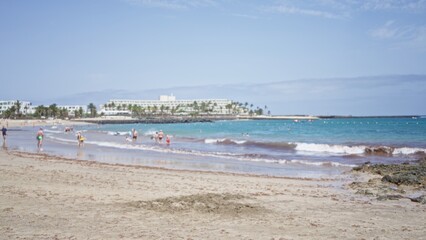 Beachgoers enjoying a sunny day at a sandy beach with clear blue water and a distant resort in the background, defocused and blurry, conveying relaxation and vacation vibes