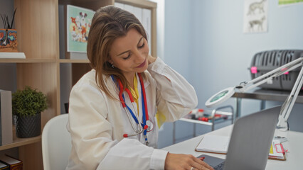 A young, attractive, blonde woman in a clinic, wearing a white coat and stethoscope, works at a...
