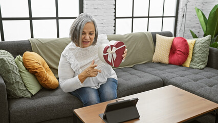 Obraz premium Mature woman sits at home on a couch smiling at a heart-shaped gift box during a video call.