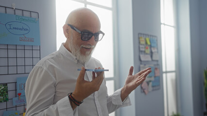 Grey-haired man with a beard using a smartphone for voice message in an indoor office setting, showcasing a modern workplace with innovation and idea boards in the background.