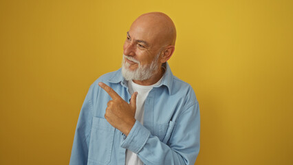 Handsome mature bald man with a grey beard and blue shirt standing isolated against a bright yellow wall while smiling and pointing to the side.