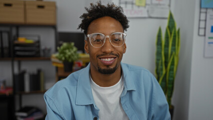 A handsome young african american man with a beard and glasses smiles while sitting in an indoor office environment.