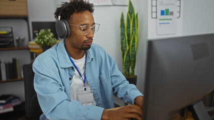 Young african american man with beard and glasses wearing headphones, working in an office at his...