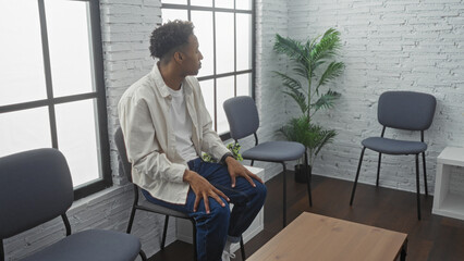 A handsome young african american man with a beard sits in an indoor waiting room, featuring a modern interior with white brick walls, windows, and blue chairs.