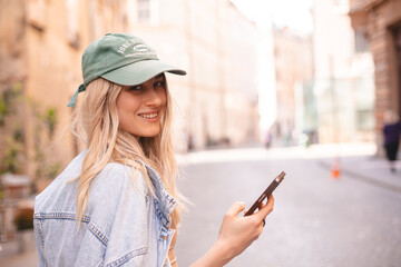 Beautiful smiling woman walking on street chatting on mobile phone. Girl talking on smartphone, texting, sending message, green cap and jeans jacket. Girl turn her head.