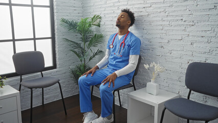 An african american male nurse in blue scrubs with a stethoscope relaxes in a hospital waiting room with white brick walls and various chairs.