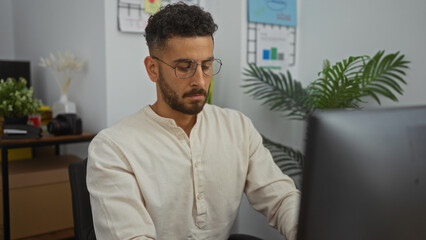 Fototapeta premium A young hispanic man wearing glasses works intently on a desktop computer in a modern office setting with plants and office decor in the background.
