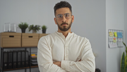 Attractive young hispanic man in a white shirt standing with arms crossed in an office environment.