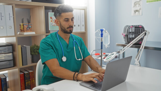 Young hispanic male veterinarian in green scrubs working on a laptop in a modern veterinary clinic office. - Powered by Adobe