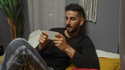 Young hispanic man in casual wear lounging on bed in a cozy bedroom, focused on his smartphone, displaying a relaxing indoor setting with home decor elements.