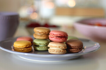 Plate of pastel macarons, cookies and chocolate, cup of tea of coffee, glass of bubble water, various berries, books and accessories on the table. Selective focus, pastel colors.
