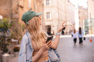 Pondering cheerful young woman with phone walking on city street checks her smartphone. Blonde girl...