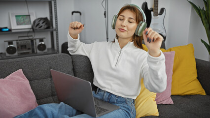 Obraz premium A joyful young woman wearing headphones dances to music while using a laptop in a cozy living room.