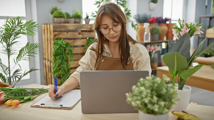 A concentrated young woman business owner in a florist shop with plants writing on clipboard laptop glasses apron.