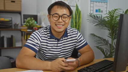 Young chinese man in a striped shirt smiling while holding a phone, seated at an office desk with a computer and plants in the background.