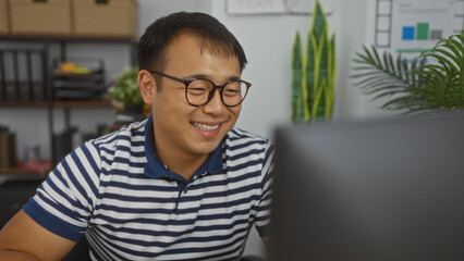 Obraz premium A young chinese man in a striped shirt and glasses smiles while working at his desk in a modern office with plants and charts in the background.