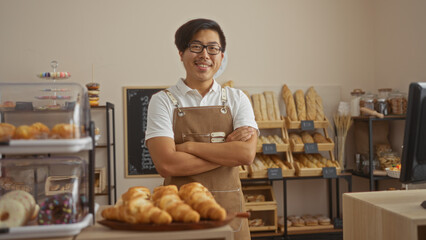 Young, chinese, man standing in a bakery with arms crossed, surrounded by fresh pastries and bread, wearing glasses and apron, looking cheerful in an indoor shop setting