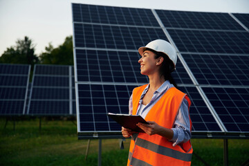 Black notepad in hands. Female worker engineer in uniform is near solar panels